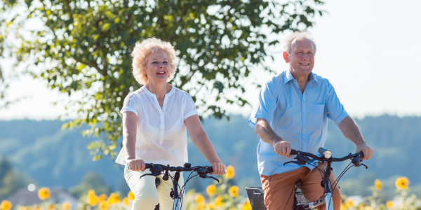 An elderly couple joyfully riding bicycles through a sunny field of sunflowers, with trees and hills in the background.