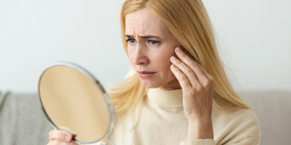 A middle‑aged blonde woman holding a small handheld mirror, touching her cheek and examining her skin with a worried expression.