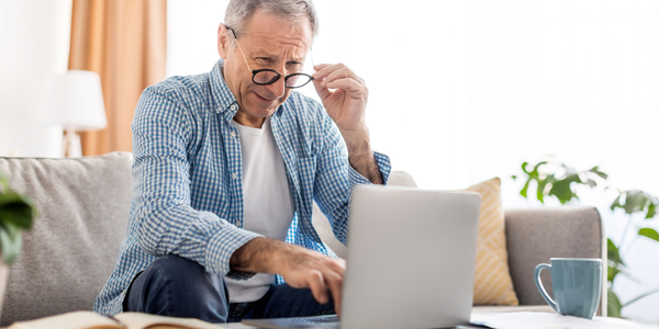 A middle-aged man sitting on a couch at home looks concerned while adjusting his glasses and using a laptop, with a coffee mug nearby.