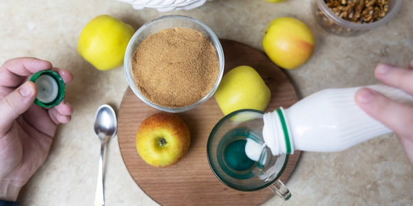 Top-down view of hands preparing a drink with apples, walnuts, and a bowl of brown powder; one hand pours a white liquid from a bottle into a glass mug while the other holds a green cap.