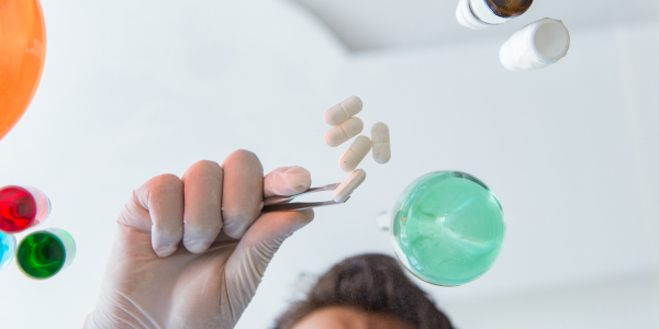 A scientist's gloved hand with tweezers holds a tablet, with more tablets on a glass table.