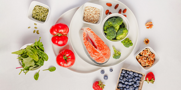 Overhead view of salmon, broccoli, leafy greens, tomatoes, berries, seeds, almonds, and walnuts arranged on white plates, illustrating nutrient-rich foods for balanced blood sugar.