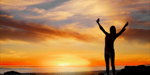 A person standing on a beach at the sunset with their arms in the air