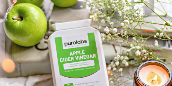 A cozy, aesthetically arranged scene featuring a Purolabs Apple Cider Vinegar supplement bottle placed on a book, surrounded by fresh green apples, delicate white flowers, and a lit candle.