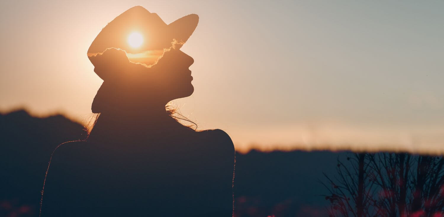 A silhouette of a woman wearing a hat with the sun in the background