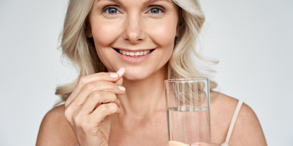 A smiling woman is holding a glass of water and a pill between her fingers.