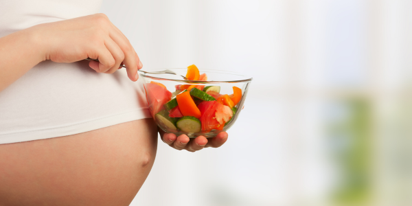 Pregnant woman in a white top holding a bowl of fresh vegetable salad with a spoon, emphasizing healthy eating during pregnancy.