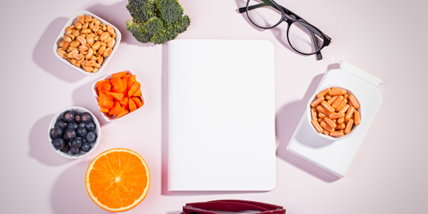  A flat lay featuring a notebook, glasses, peanuts, carrots, blueberries, broccoli, orange slices, and a container of orange capsules on a pink background.