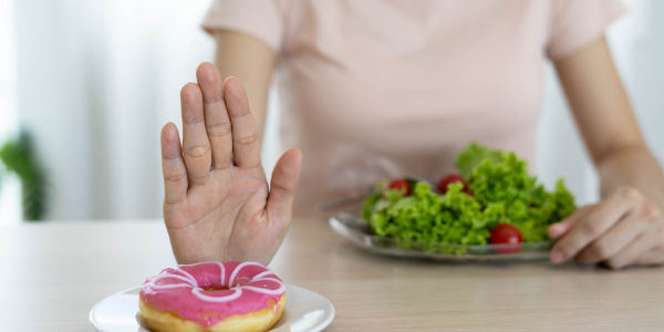 Hand gesturing "stop" to a plate with a pink frosted doughnut and green salad, illustrating avoiding ultra-processed junk foods and selecting healthier whole food options.