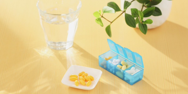 A glass of water, a white dish with yellow capsules, and a blue weekly pill organizer containing various pills, all placed on a light wooden surface with a green plant in the background.