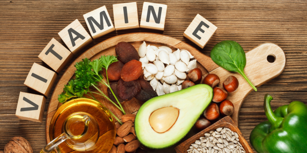 A wooden board displaying various foods rich in vitamin E, including avocado, spinach, green bell pepper, and a small jar of vegetable oil. Wooden letter blocks spell out “VITAMIN E” at the top.