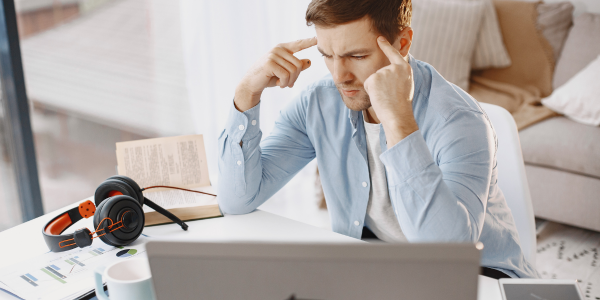 A young man sits at a desk in a bright living-room setting, leaning toward his open laptop with his fingers pressed to his temples in frustration.