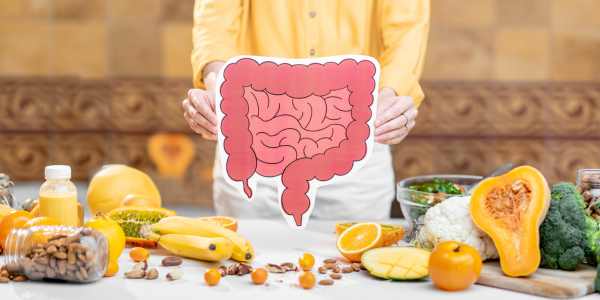 Person in a yellow shirt holds a stylized illustration of the colon above a table covered with high-fiber foods like bananas, nuts, oranges, broccoli, and squash.