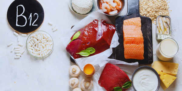 Assortment of vitamin B12-rich foods and supplements, including salmon, liver, beef, cheese, yogurt, milk, eggs, shrimp, canned fish, oats, mushrooms, and a bowl of white capsules next to a chalkboard labeled 'B12.