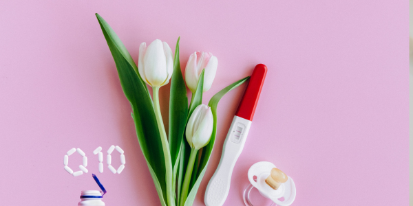 White tulips, a pregnancy test, a pacifier, and Q10 capsules arranged on a pink background.