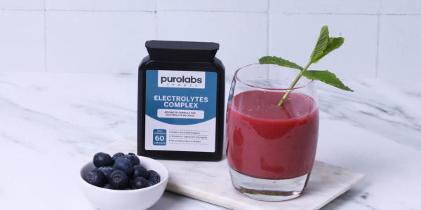Purolabs “Electrolytes Complex” bottle beside a red berry smoothie garnished with mint and a small bowl of blueberries on a marble surface.
