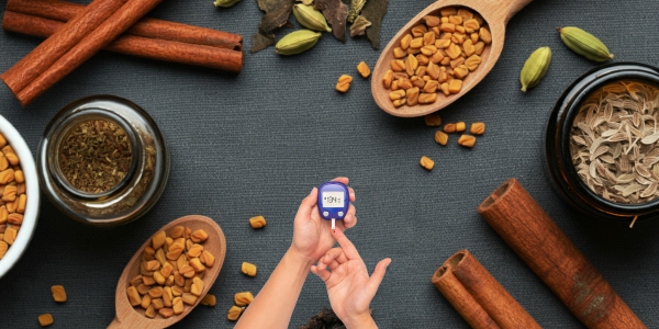 Hand using a blood glucose meter surrounded by cinnamon, fenugreek seeds and herbs.