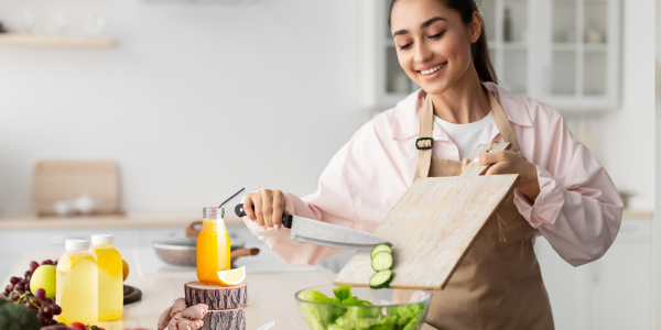 Smiling woman in an apron preparing a fresh salad, sliding sliced cucumbers from a cutting board into a glass bowl in a bright modern kitchen.