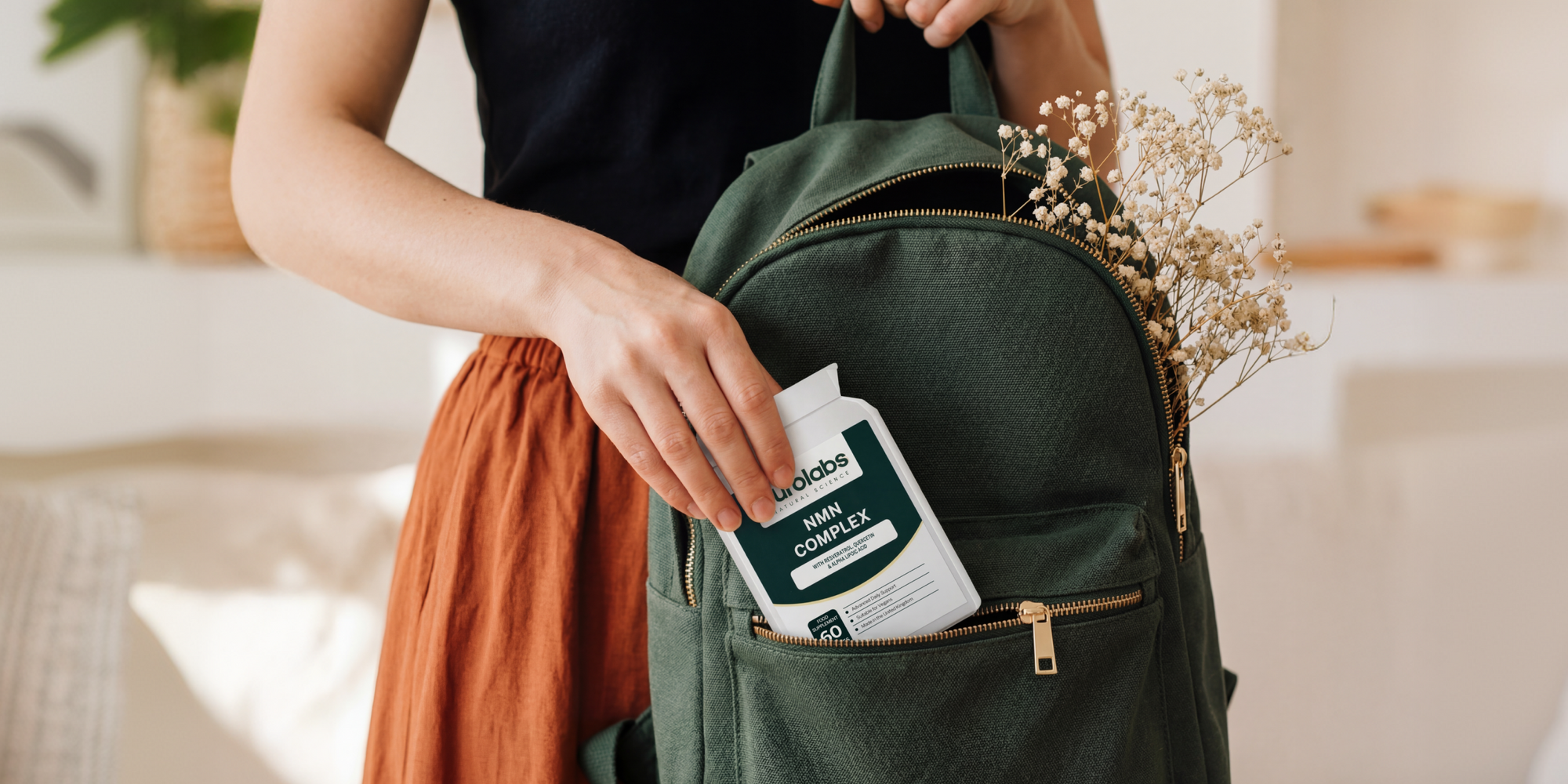 A person places a bottle of NMN Complex supplements into the front pocket of a dark green backpack, with small dried flowers tucked into the bag’s side.