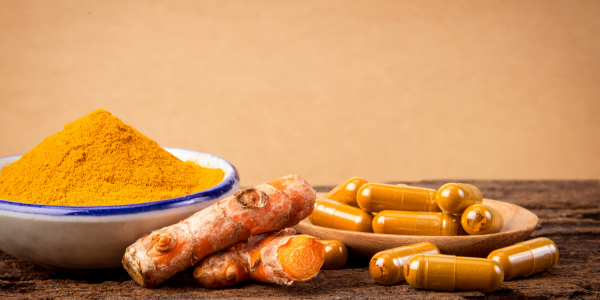 A bowl of bright turmeric powder beside fresh turmeric roots and turmeric supplement capsules on a wooden surface.